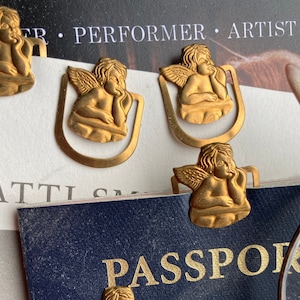 May include: Several gold-coloured angel-shaped metal bookmarks. The bookmarks are attached to a blue passport and a white card with the word "PERFORMER" visible. The angels are in a seated pose with their hands on their chins.