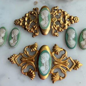 May include: An assortment of antique gold-toned metal jewelry findings. The collection includes ornate filigree pieces, oval cameos with a woman's profile in white against a green background, and decorative clasps. The pieces are arranged on a light-colored surface.