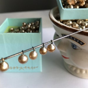 May include: A close-up of a string of pearl beads hanging from a metal rod. The beads are a light gold color and are arranged in a row. The beads are hanging over a light blue box with a gold design. The box is filled with more pearl beads.