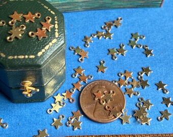 以下が含まれることがあります： 青い表面に散らばった小さな金色の金属製星型チャームのコレクション。緑色の八角形の箱と銅貨の隣にあります。下部には木製の定規が見え、背景には2匹の魚が描かれた長方形のプリントが配置されています。
