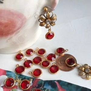 May include: A collection of red gemstone charms with gold-colored settings. One earring features a gold-colored flower design with a clear rhinestone center and a red gemstone drop. The charms are scattered on a white surface.