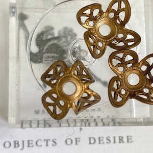 May include: Close-up of several small, gold-colored metal bead caps with a floral design. The bead caps are inside a clear, square container. The text "OBJECTS OF DESIRE" is visible at the bottom of the image.