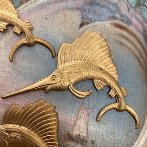 Może przedstawiać: Trzy złote metalowe ozdoby w kształcie ryb z detalicznym wzorem. Ryby są ułożone w grupie na teksturowanym, niebiesko-brązowym tle.