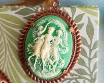 May include: A green cameo pendant with a copper-colored frame. The cameo features a white relief of three figures. The pendant is oval-shaped and has a twisted rope-like border. The background is a patterned box with a ribbon.