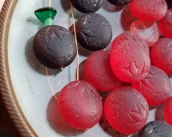 May include: A collection of red and black glass beads with a leaf design. The beads are arranged on a white plate with a gold rim.