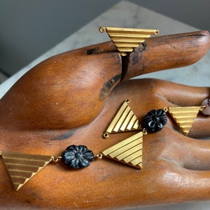 以下が含まれることがあります： 黒い花の形をしたビーズと三角形のペンダントチャームが付いた金のネックレス。ネックレスは木製の手に置かれています。