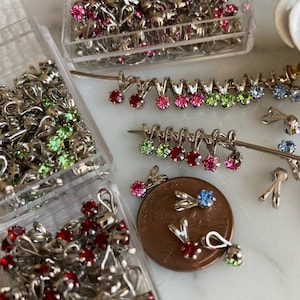 May include: Assortment of silver-toned metal jewelry findings with colorful rhinestones. The findings are in various shapes and sizes, including loops, teardrops, and round shapes. The rhinestones are red, green, blue, and pink.