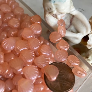May include: A close-up of a large quantity of pink iridescent shell-shaped beads. The beads are arranged in a clear plastic container. A US penny is included for scale.