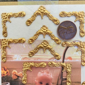 Puede incluir: Un conjunto de adornos de esquina de metal de color dorado con un diseño floral. Los adornos están dispuestos sobre una superficie blanca con un centavo para la escala.