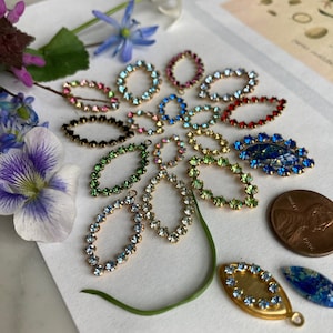 May include: Assortment of colorful oval-shaped rhinestone embellishments with gold settings. The rhinestones are arranged in a fan shape on a white surface.