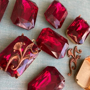 May include: A collection of seven red glass cabochons, some with ornate metal accents. The cabochons are rectangular and have a faceted cut. They are arranged on a blue textured surface.