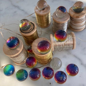 Peut inclure: Une collection de billes de verre iris&eacute;es dispos&eacute;es sur des bobines de fil. Les billes sont dispos&eacute;es en cercle autour des bobines, certaines &eacute;tant plac&eacute;es sur les bobines. Les bobines sont en bois et de diff&eacute;rentes couleurs, notamment or, argent et blanc.