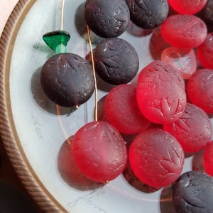 May include: A collection of red and black glass beads with a leaf design. The beads are arranged on a white plate with a gold rim.