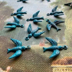 Puede incluir: Una colección de abalorios de pájaros de metal azul turquesa. Los abalorios tienen forma de pájaro volando con un pequeño bucle en la parte superior para sujetarlos a un collar, pulsera u otras joyas.