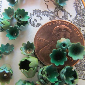 May include: A collection of green metal flower embellishments, some with a copper center, scattered on a white surface with a copper penny featuring the words "In God We Trust" and "Liberty".