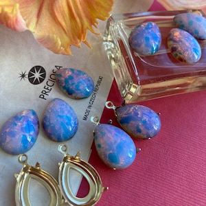 Könnte beinhalten: Sortiment aus irisierenden blauen und rosa gläsernen Tropfenperlen mit goldenen Metallfassungen. Einige Perlen sind lose und einige sind an den Fassungen befestigt. Die Perlen befinden sich auf einer weißen Oberfläche mit dem Text "PRECOSA MADE IN CZECH REPUBLIC".