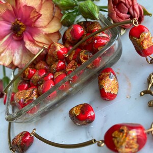 May include: A gold bracelet with red and gold glass beads. The beads have a speckled, textured surface. The bracelet is lying on a white surface with a few beads scattered around it.