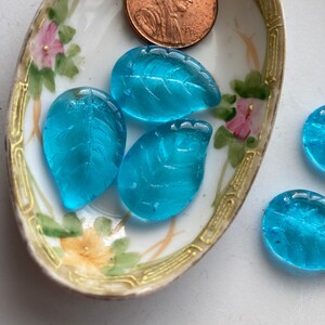 May include: An oval, antique porcelain dish with floral designs holds three blue glass leaf beads and a copper penny. The dish has a gold trim and painted flowers in pink and yellow. The leaf beads are a vibrant turquoise color.