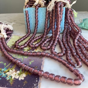 May include: A vintage box filled with strands of purple glass beads. The box is blue and has a label that reads "ST 08 Amy".