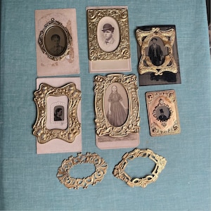 Puede incluir: Una colección de ocho marcos de fotos antiguos de metal dorado adornado con inserciones de vidrio. Los marcos están dispuestos sobre una superficie azul claro. Algunos de los marcos contienen fotografías en blanco y negro de personas.