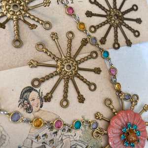 May include: A gold necklace with a variety of colorful glass beads and star-shaped charms. The necklace is on a vintage background with a woman's image and the number "152".