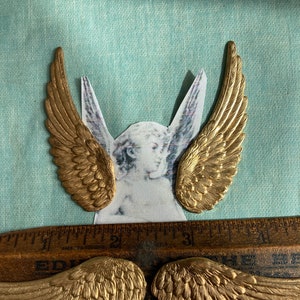 May include: Four gold-colored metal angel wings. The wings are detailed with a textured feather design. The wings are arranged in two pairs, with one pair on top of the other.