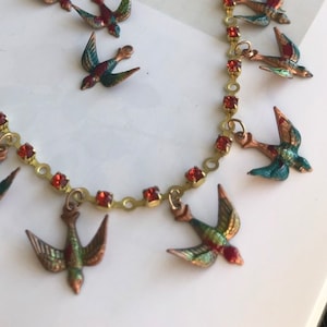 Può includere: Una collana a catena dorata con accenti di cristallo rosso e ciondoli di uccelli smaltati pendenti. Gli uccelli sono multicolori con tonalità verdi, blu e marroni.