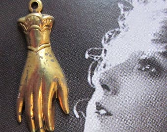 May include: A gold-toned metal charm in the shape of a hand with a detailed design. The charm has a loop at the top for attaching to a necklace or bracelet.
