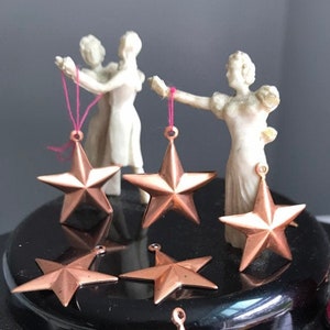 May include: A collection of eight copper five-pointed stars with a pink thread and two white figurines holding the stars.