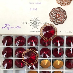 Puede incluir: Una caja de cristales Rivoli Siam, de 18 mm, con engastes de filigrana dorados. La caja está etiquetada con texto que incluye "Secunda", "Art. 1122" y "Made in Austria". Los cristales son de color rojo intenso.