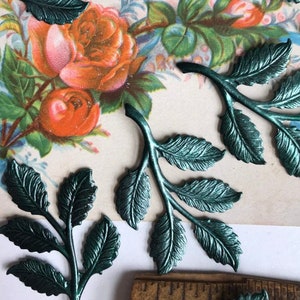 May include: A set of six teal green metal leaf sprigs. The sprigs are made of thin metal and have a slightly textured surface. The leaves are arranged in a cluster, with some overlapping. The sprigs are approximately 2 inches in length.