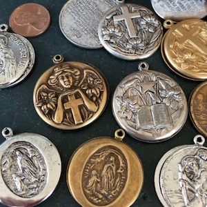 May include: A collection of round metal religious pendants in silver and gold tones. The pendants feature embossed designs including crosses, angels, open books, and figures of saints. Some pieces display engraved text such as the Serenity Prayer.