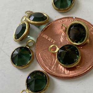 May include: Close-up of several dark green gemstone pendants with gold-colored bezels and attached loops. The pendants are various shapes, including round and oval. A copper penny is next to the pendants.
