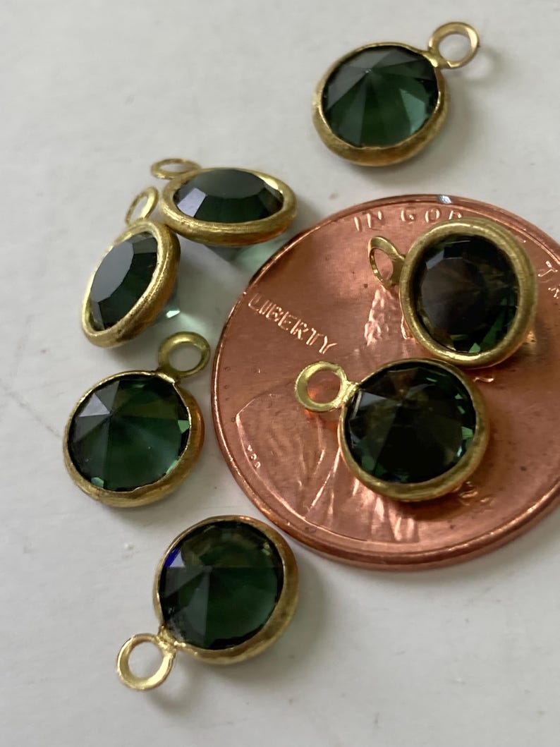 May include: Close-up of several dark green gemstone pendants with gold-colored bezels and attached loops. The pendants are various shapes, including round and oval. A copper penny is next to the pendants.