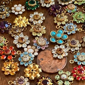 May include: A selection of vintage flower-shaped jewellery components in gold, red, green, blue, and purple. The components are decorated with rhinestones and gemstones, and are arranged on a wooden surface.