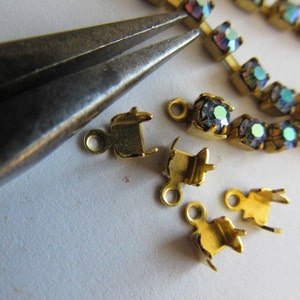 Puede incluir: Primer plano de cierres de metal dorado para la elaboración de joyas. Los cierres son pequeños y tienen un diseño sencillo. También se puede ver un par de alicates en la imagen.