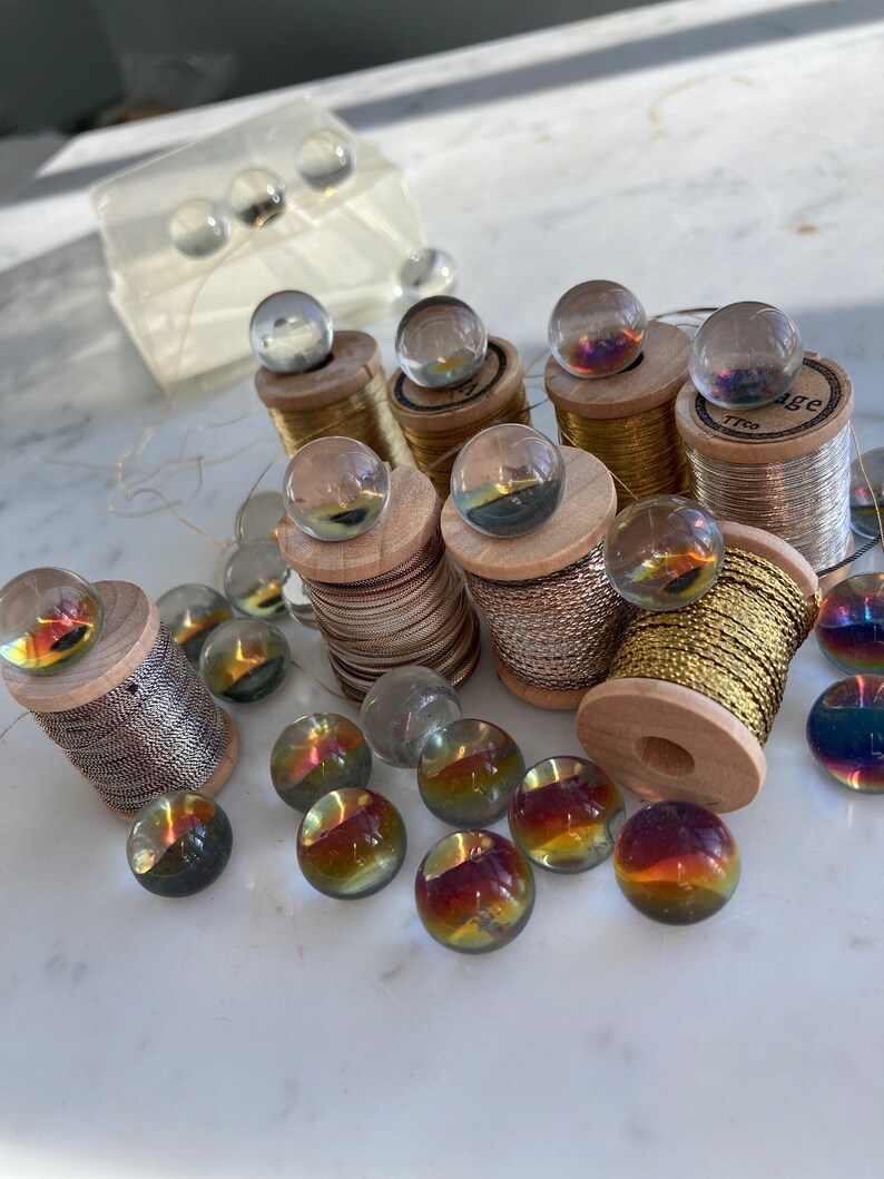 Peut inclure: Une collection de billes en verre iris&eacute;es et de bobines de fil d'or et d'argent. Les billes sont dispos&eacute;es sur une surface en marbre blanc. Les bobines de fil sont sur des bobines en bois avec des &eacute;tiquettes.