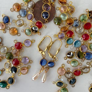 May include: An assortment of colorful, oval-shaped gemstone charms with gold-colored metal settings. Two gold-colored earrings with blue gemstones and pearl drops are also visible. The charms are in various colors, including blue, green, red, and orange.