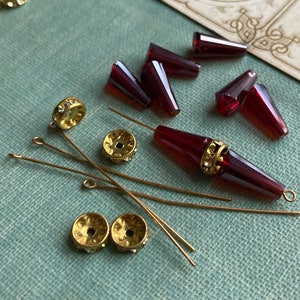 May include: Assortment of red glass beads, gold-toned metal beads, and gold-toned head pins on a teal fabric surface.