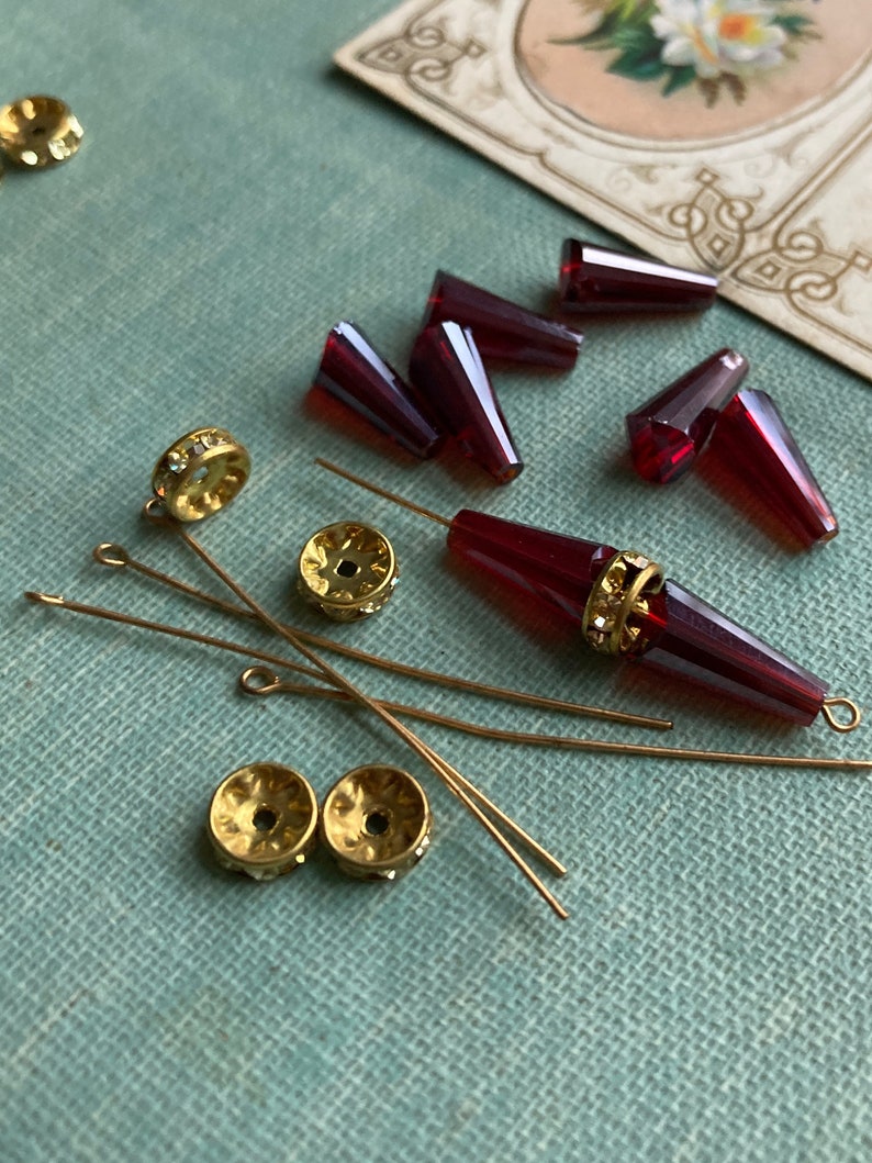 May include: Assortment of red glass beads, gold-toned metal beads, and gold-toned head pins on a teal fabric surface.