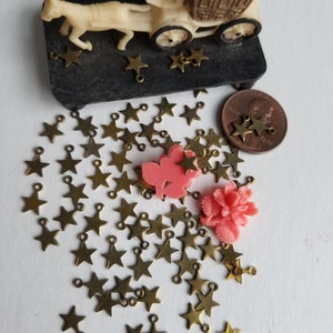 K&ouml;nnte beinhalten: Eine gro&szlig;e Menge goldener sternf&ouml;rmiger Charms mit Sprungringen. Die Charms sind auf einer wei&szlig;en Oberfl&auml;che verstreut. Es befinden sich zwei rosa Plastikblumen im Bild.
