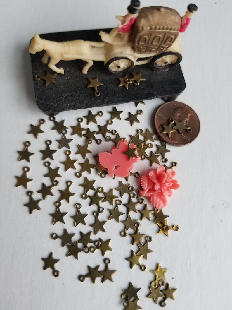 K&ouml;nnte beinhalten: Eine gro&szlig;e Menge goldener sternf&ouml;rmiger Charms mit Sprungringen. Die Charms sind auf einer wei&szlig;en Oberfl&auml;che verstreut. Es befinden sich zwei rosa Plastikblumen im Bild.