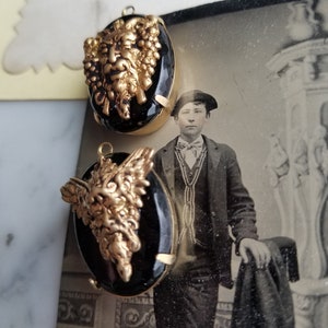 May include: Two gold-toned pendant charms with black glass cabochons. Each charm features a detailed, three-dimensional, gold-toned face with a beard and grapes.
