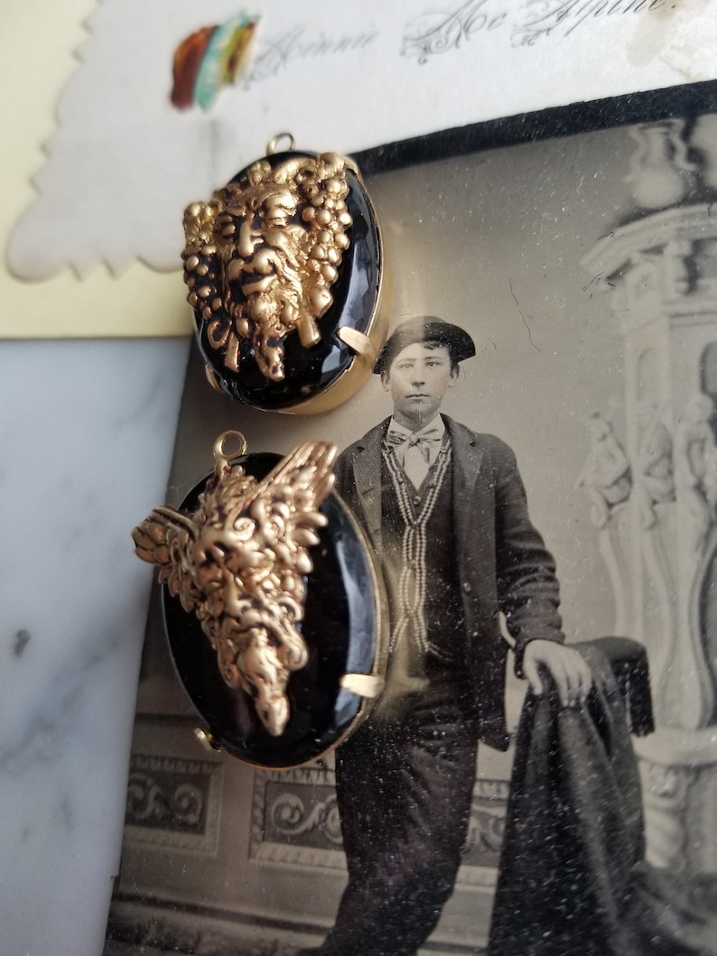 May include: Two gold-toned pendant charms with black glass cabochons. Each charm features a detailed, three-dimensional, gold-toned face with a beard and grapes.