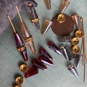 May include: Assortment of glass beads in various colours and shapes, including clear, amber, and red. Some beads have gold accents. The beads are arranged on a textured surface with a US penny for scale.
