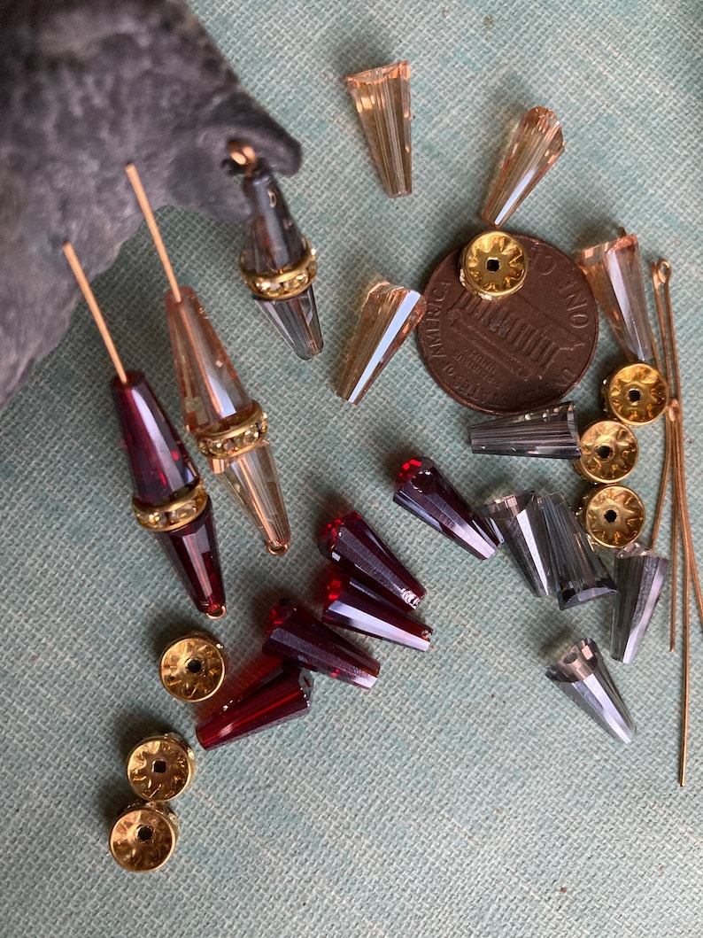 May include: Assortment of glass beads in various colours and shapes, including clear, amber, and red. Some beads have gold accents. The beads are arranged on a textured surface with a US penny for scale.