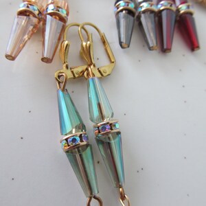 May include: A collection of dangle earrings in various colors, including iridescent teal, rose, gray, and burgundy. Each earring features a conical crystal pendant with a gold-colored band adorned with iridescent rhinestones. The earrings are suspended from gold-colored lever-back clasps.