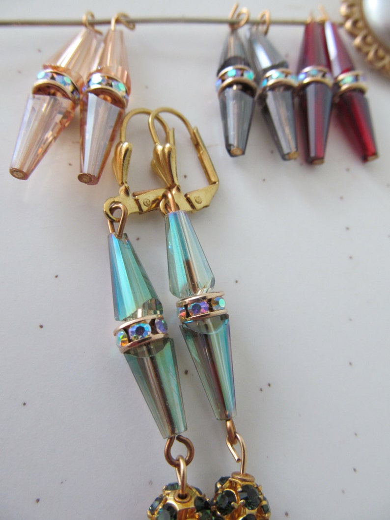 May include: A collection of dangle earrings in various colors, including iridescent teal, rose, gray, and burgundy. Each earring features a conical crystal pendant with a gold-colored band adorned with iridescent rhinestones. The earrings are suspended from gold-colored lever-back clasps.