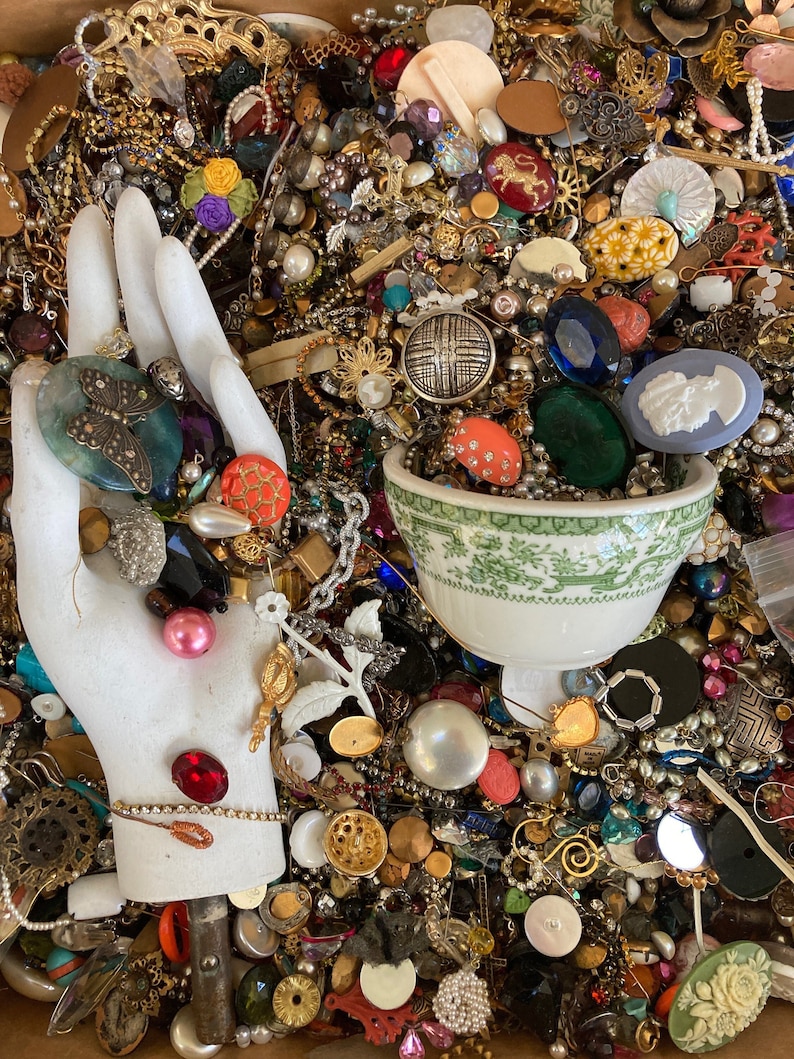 May include: A large collection of colorful buttons, beads, and jewelry findings in a box. The items are arranged in a random pattern and include a variety of materials, such as metal, glass, plastic, and wood.
