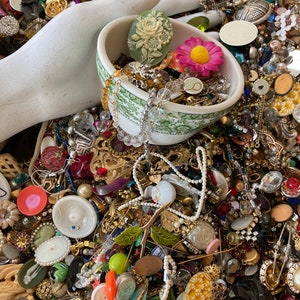 May include: A white ceramic bowl filled with a variety of colorful buttons, beads, and jewelry. The bowl is sitting on a table covered in a large collection of buttons, beads, and jewelry.